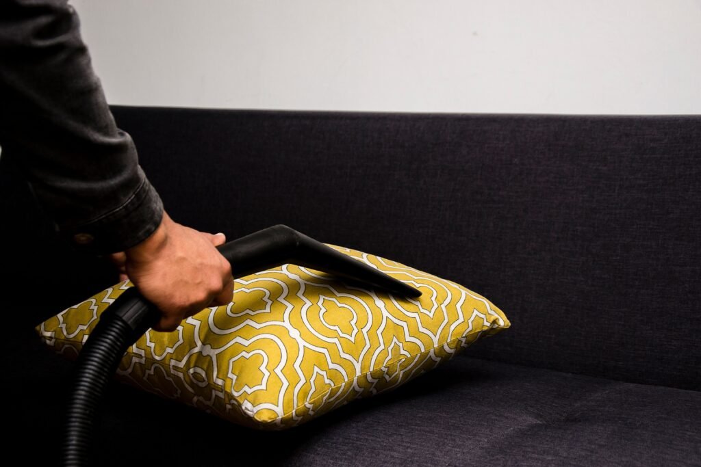 أفضل شركة تنظيف مجالس في أبوظبي0556485364 Close-up of a person vacuuming a patterned pillow on a dark sofa indoors.