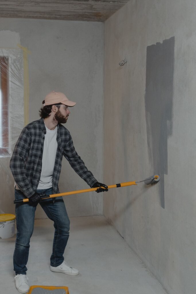 أفضل شركة صيانة وترميم جدران بالعين 0547761290 An adult man wearing a hat and gloves is painting an indoor wall with a roller.